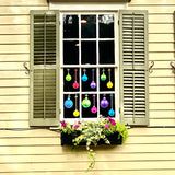 Green shutters frame a window displaying Cover-Alls Christmas Ornament Ball Decals, as a flower box with lush plants sits below.
