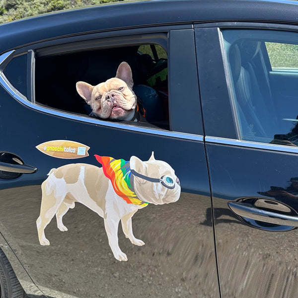 A French Bulldog peeks out of the rear window of a black car, which has a large decal featuring a Cover-Alls Custom Pet Drawing of a similar-looking dog wearing goggles and a rainbow-colored cape.