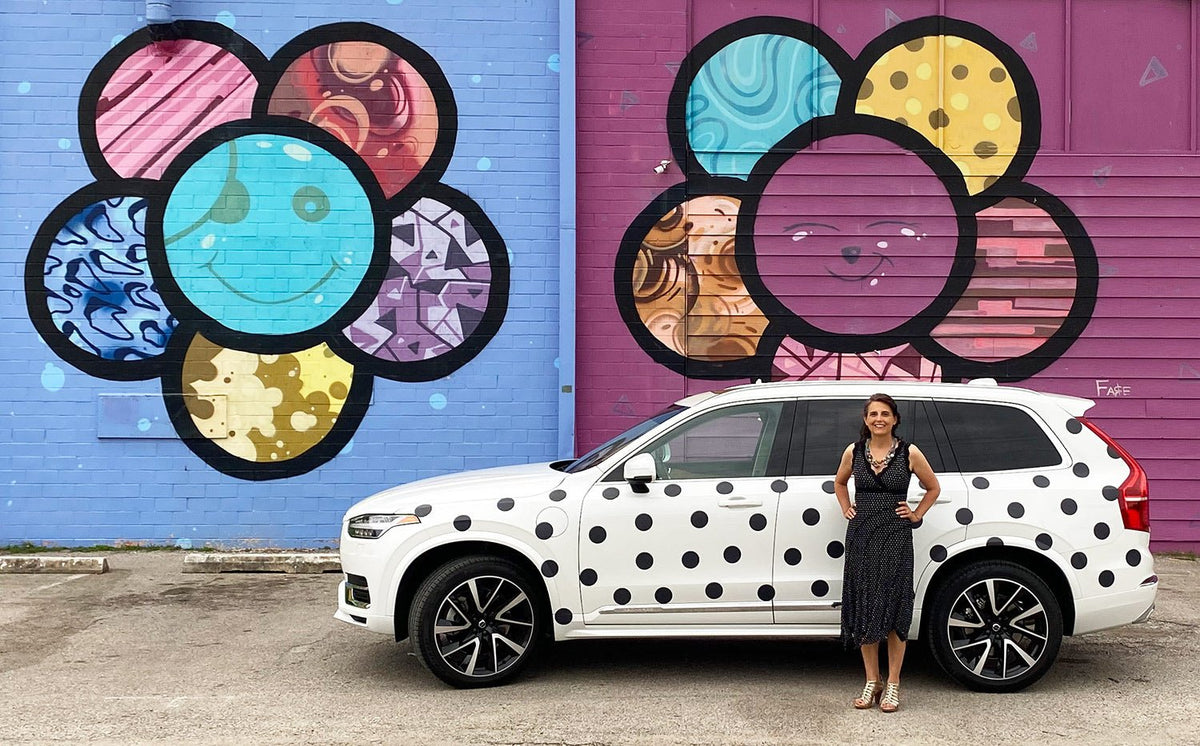 A woman standing beside a white SUV parked in front of a vibrant mural featuring large, colorful Cover-Alls Dot Decals on a purple wall.