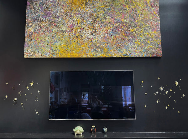 A colorful abstract painting above a television, mounted on a black wall decorated with Cover-Alls Gold Twinkly Stars, with small figurines on a shelf below.