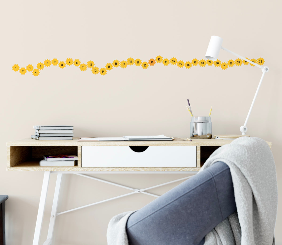 A neat wooden desk with books and a pen holder sits against a wall adorned with Cover-Alls Create a New Habit Daily Cog Stickers and a border of yellow sunflowers. A gray blanket is draped over a white chair.