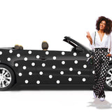A woman with curly hair, standing by a black convertible car with Cover-Alls white polka dot decals, is smiling and waving. She wears a white blouse and polka-dotted pants.