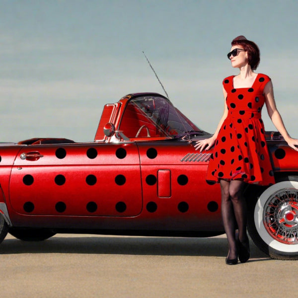 A woman in a red polka dot dress and sunglasses leaning on a red car with Cover-Alls Dot Decals, standing in front of a building.