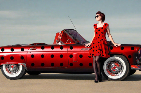 A woman in a red polka dot dress and sunglasses leaning on a red car with Cover-Alls Dot Decals, standing in front of a building.