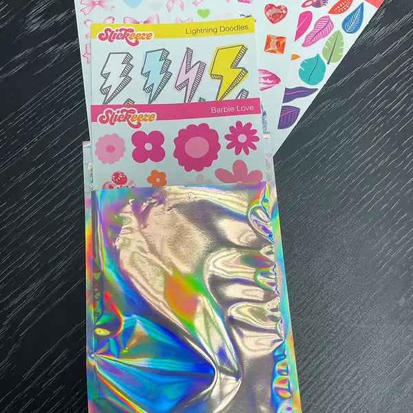 Four packs of Stickeeze Kid's Sticker Subscription Club stickers with various designs, including gems, bows, lightning doodles, and flowers, made from easy-peel material, lie on a black surface next to a partially opened holographic envelope.