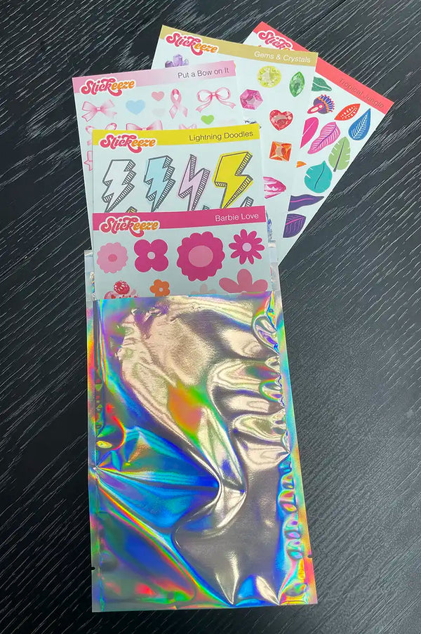Four packs of Stickeeze Kid's Sticker Subscription Club stickers with various designs, including gems, bows, lightning doodles, and flowers, made from easy-peel material, lie on a black surface next to a partially opened holographic envelope.