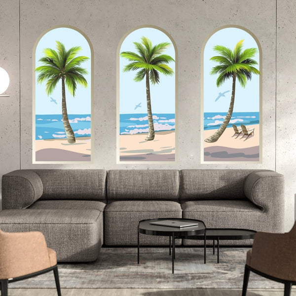 A modern living room with a gray sofa and two brown chairs facing each other, three framed beach wall decor paintings above the sofa, a round lamp, and a set of black coffee tables featuring Palm Tree Arch Scene Decals by Cover-Alls.