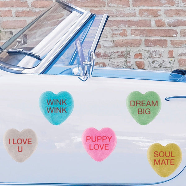 A vintage blue convertible car with heart-shaped stickers on the windshield displaying phrases like "wink wink" and "dream big" against a brick wall background, adorned with Cover-Alls candy hearts for Valentine's Day.