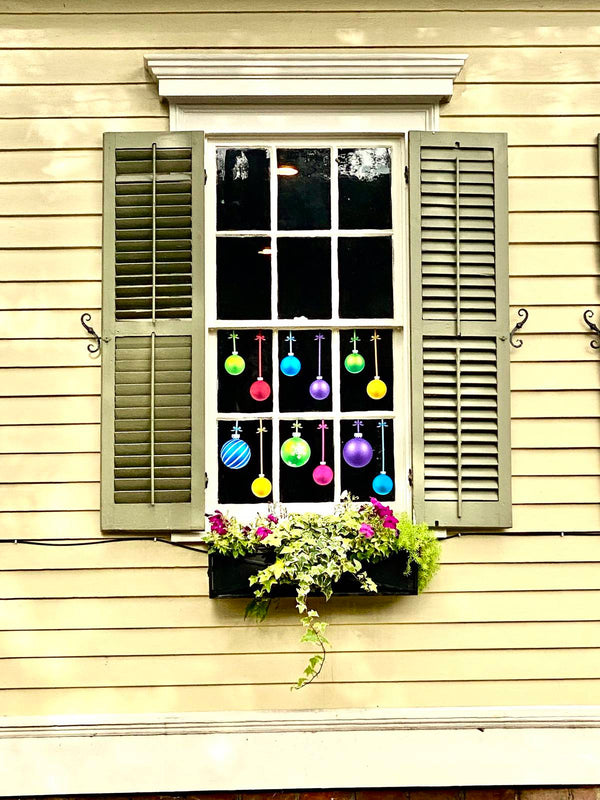 Green shutters frame a window displaying Cover-Alls Christmas Ornament Ball Decals, as a flower box with lush plants sits below.