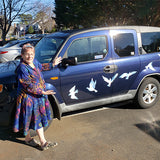 A person stands beside a blue SUV decorated with Cover-Alls' Birds of Peace Decals, featuring white watercolor-like birds in flight.