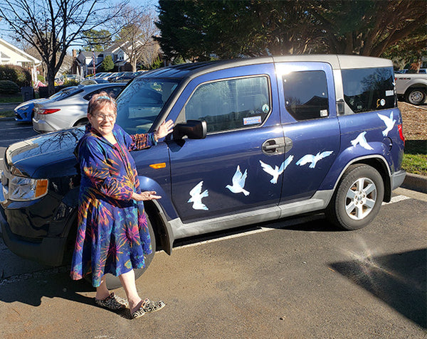 A person stands beside a blue SUV decorated with Cover-Alls' Birds of Peace Decals, featuring white watercolor-like birds in flight.