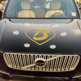 Front view of a black Volvo car adorned with a Gold Glitter #13 in Gold Heart decoration by Cover-Alls on the hood, parked in front of various buildings. Perfect for a Swiftie planning their next concert road trip.