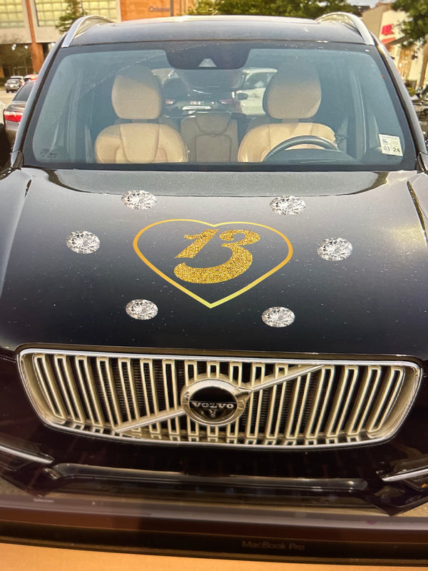 Front view of a black Volvo car adorned with a Gold Glitter #13 in Gold Heart decoration by Cover-Alls on the hood, parked in front of various buildings. Perfect for a Swiftie planning their next concert road trip.