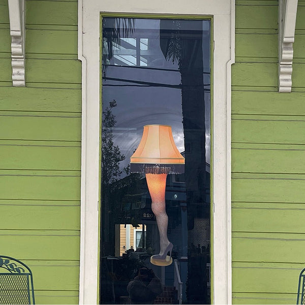 A "A Christmas Story's Leg Lamp" decal with a fringed shade displayed in a narrow window of a green house, reflecting trees and a cloudy sky.