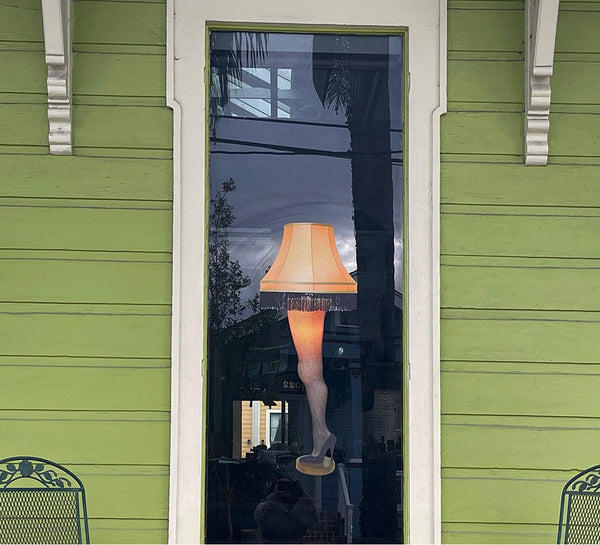 A "A Christmas Story's Leg Lamp" decal with a fringed shade displayed in a narrow window of a green house, reflecting trees and a cloudy sky.