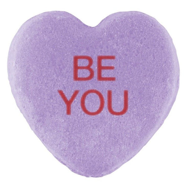 A purple heart-shaped Candy Hearts for Pride with the phrase "be you" printed in red lettering on its three-dimensional surface by Cover-Alls.
