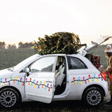 A person dressed in a winter coat packs gifts into a compact white car decorated with Cover-Alls' Christmas Lights Decals, spreading holiday cheer. A tree is securely fastened atop the car.