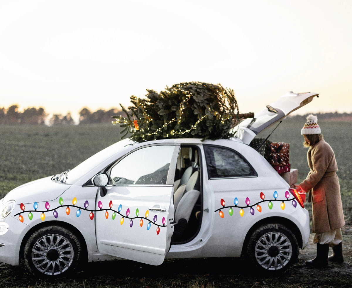 A person dressed in a winter coat packs gifts into a compact white car decorated with Cover-Alls' Christmas Lights Decals, spreading holiday cheer. A tree is securely fastened atop the car.