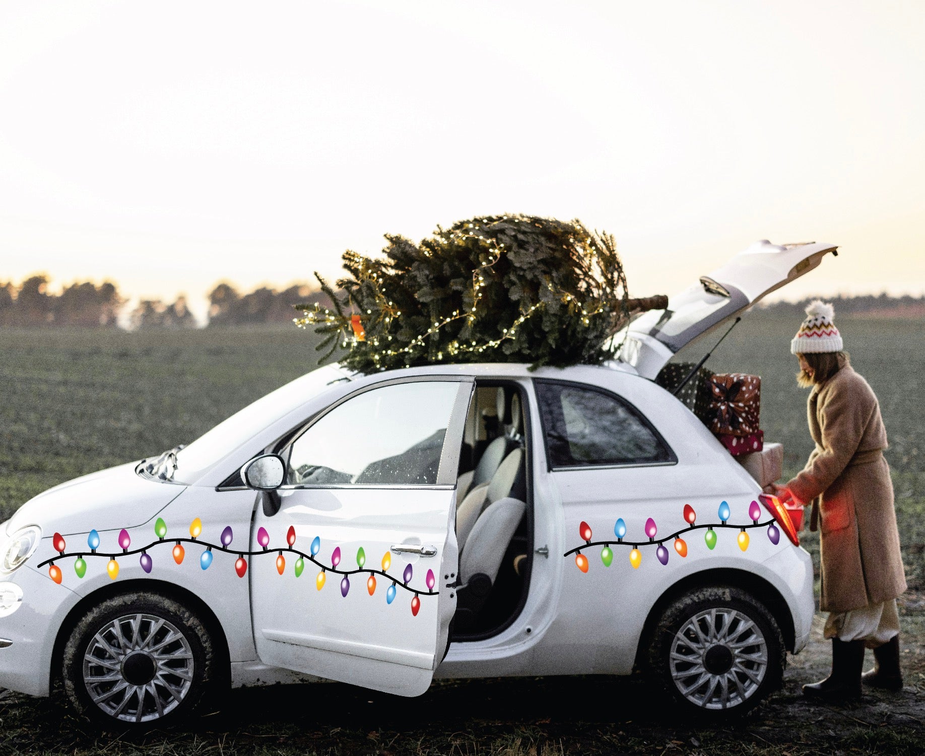  A person dressed in a winter coat packs gifts into a compact white car decorated with Cover-Alls' Christmas Lights Decals, spreading holiday cheer. A tree is securely fastened atop the car.