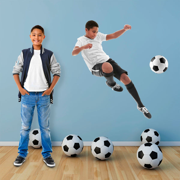 A boy stands smiling, next to a Cover-Alls Custom Photo Cutout of himself performing a mid-air soccer kick, surrounded by multiple soccer balls.
