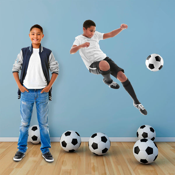 A boy stands smiling, next to a Cover-Alls Custom Photo Cutout of himself performing a mid-air soccer kick, surrounded by multiple soccer balls.