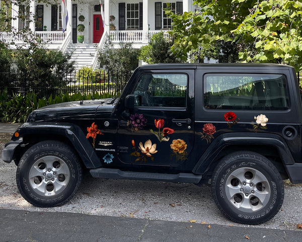 A black Jeep Wrangler parked on a gravel path with Cover-Alls Dark Romance Vintage Flower Decals on its side, in front of a row of white houses with porches and greenery.