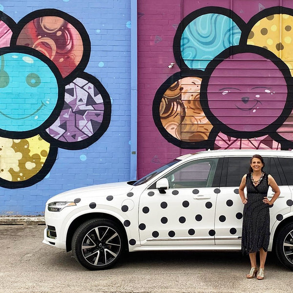 A woman standing beside a white SUV parked in front of a vibrant mural featuring large, colorful Cover-Alls Dot Decals on a purple wall.