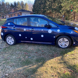 A blue compact car with large Cover-Alls white polka dot decals parked on grass, with trees and a brick building in the background.
