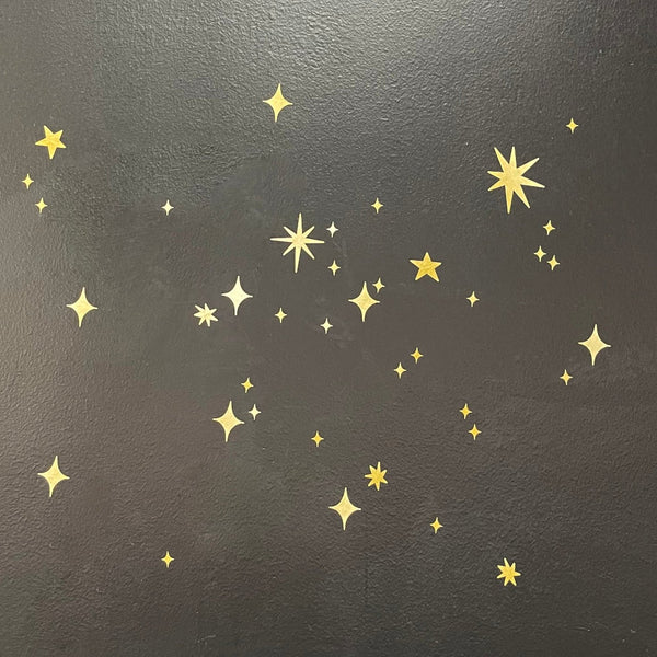 Gold Twinkly Stars stickers of various sizes scattered randomly on a dark gray background, creating a magical scene decor. (Cover-Alls)
