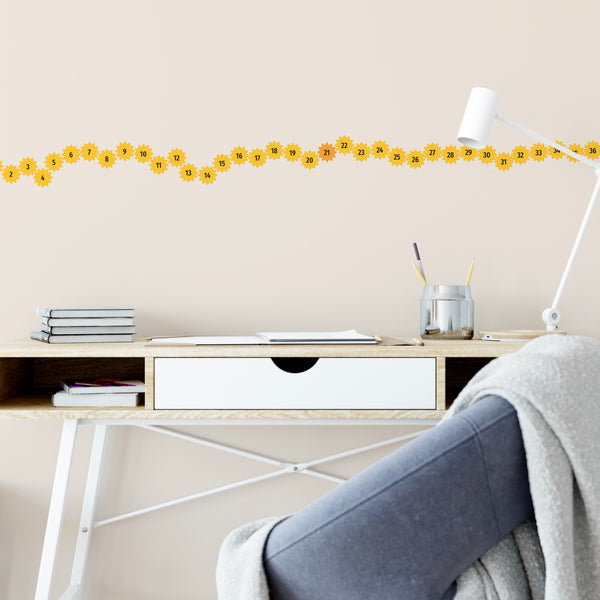 A neat wooden desk with books and a pen holder sits against a wall adorned with Cover-Alls Create a New Habit Daily Cog Stickers and a border of yellow sunflowers. A gray blanket is draped over a white chair.