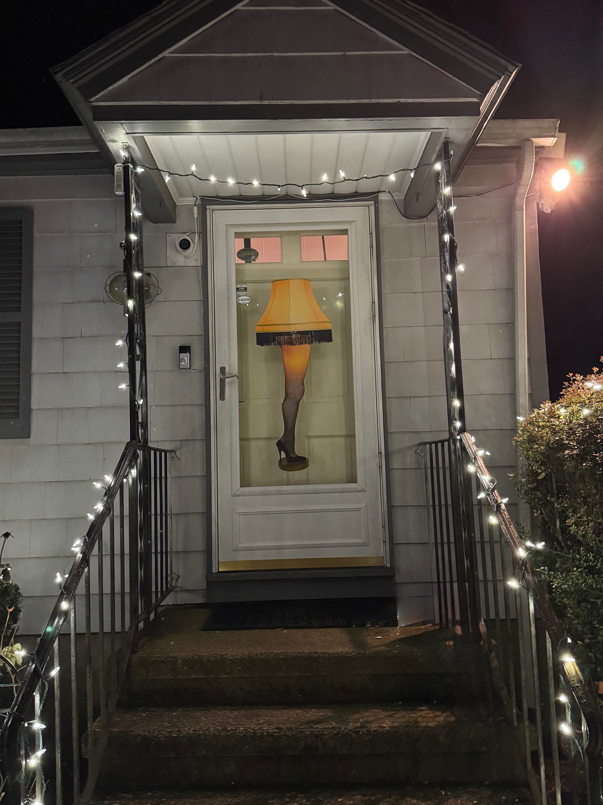 The front door features "A Christmas Story's Leg Lamp" by Cover-Alls, adorned with a fringed shade for classic Christmas humor. Adhesive cloth decals and string lights add to the porch's quirky charm.