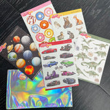 Five sheets of assorted reusable stickers from Stickeeze's Kid's Sticker Club on a dark wooden surface, including designs of planets, doughnuts, spring animals, race cars, and dinosaurs, on two iridescent envelopes.