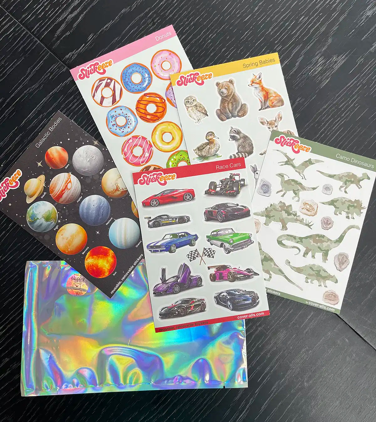 Five sheets of assorted reusable stickers from Stickeeze's Kid's Sticker Club on a dark wooden surface, including designs of planets, doughnuts, spring animals, race cars, and dinosaurs, on two iridescent envelopes.