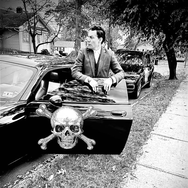 A man in a suit exiting a black car adorned with Skull & Crossbone Decals on the front grille, parked on a suburban street.