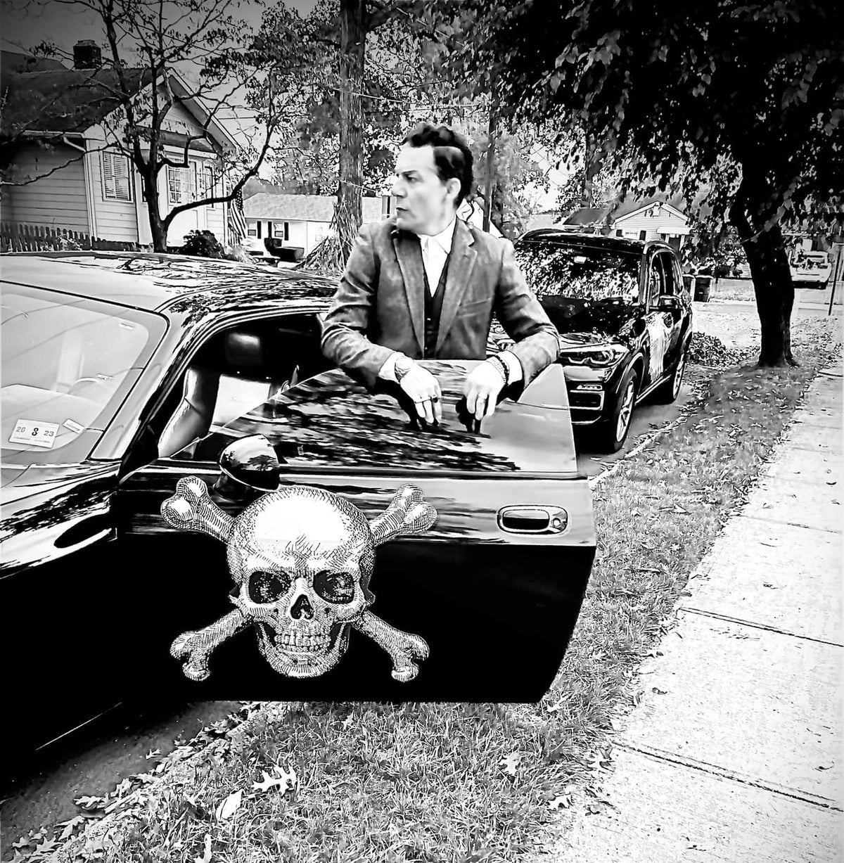 A man in a suit exiting a black car adorned with Skull & Crossbone Decals on the front grille, parked on a suburban street.