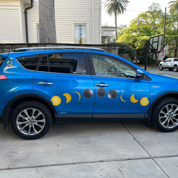 A blue car with Cover-Alls Total Solar Eclipse Decals on its side, parked beside a residential street with trees and houses in the background.