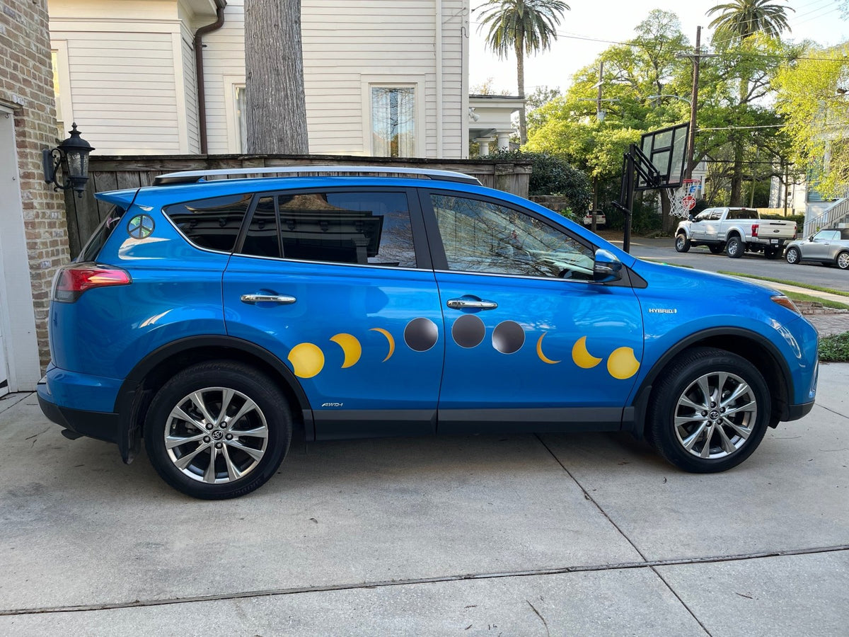 A blue car with Cover-Alls Total Solar Eclipse Decals on its side, parked beside a residential street with trees and houses in the background.