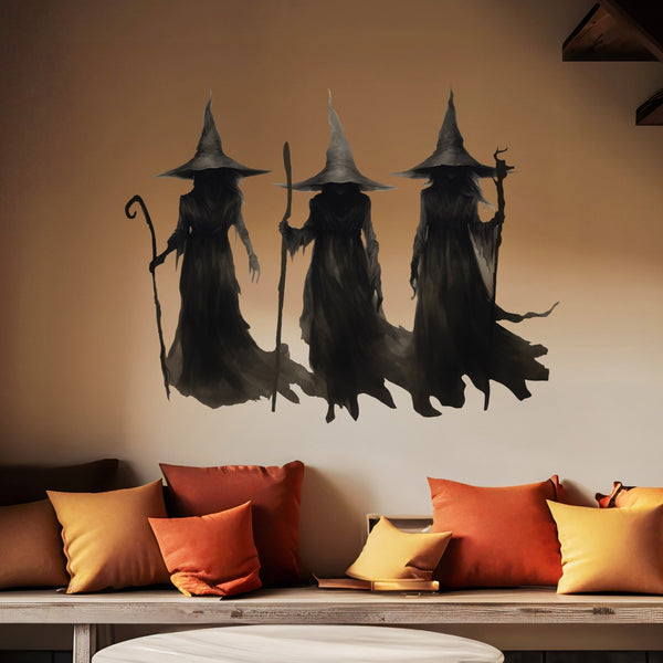 Three figures in dark, wizard-like robes and pointed hats, part of the Cover-Alls' "A Trio of Witches" collection, are painted on the wall above a couch adorned with orange and yellow pillows, creating hauntingly beautiful witch-themed wall decor.