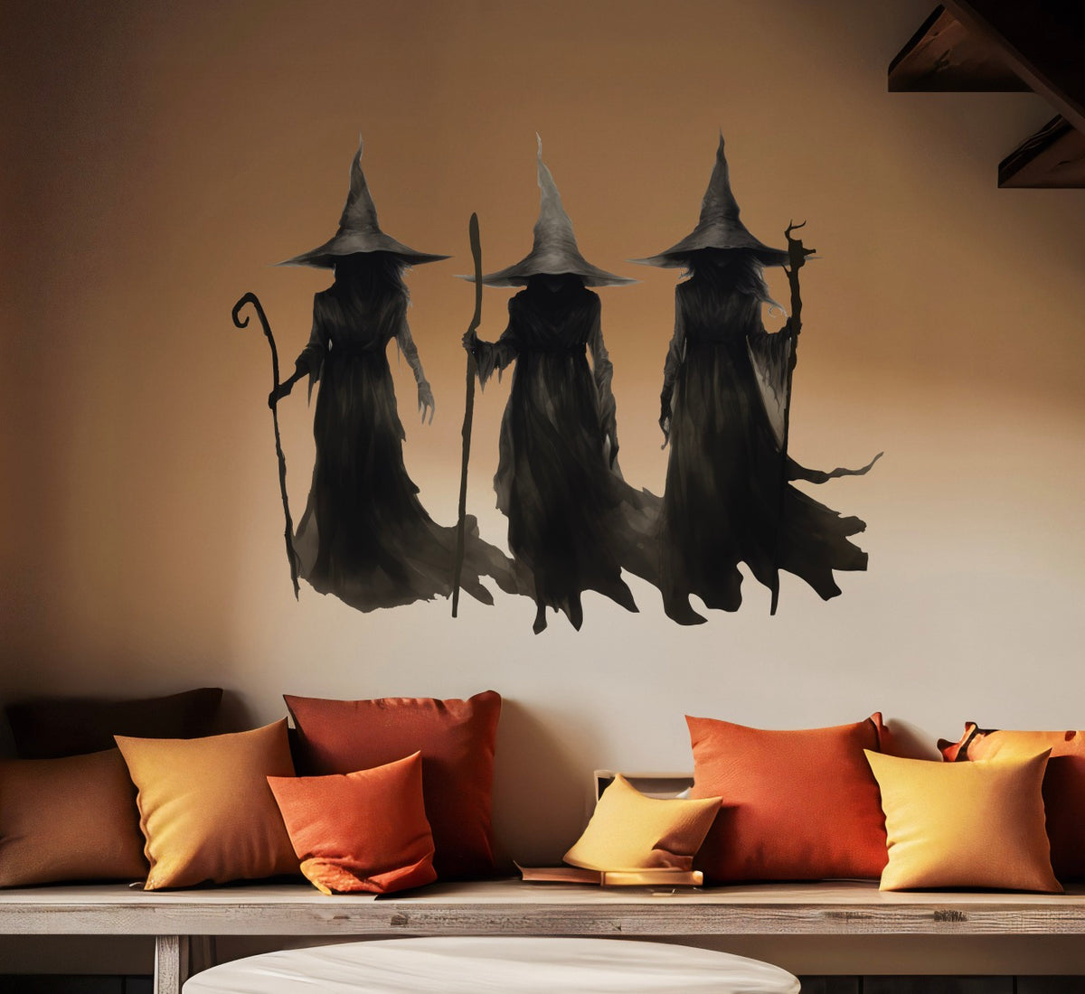 Three figures in dark, wizard-like robes and pointed hats, part of the Cover-Alls' "A Trio of Witches" collection, are painted on the wall above a couch adorned with orange and yellow pillows, creating hauntingly beautiful witch-themed wall decor.