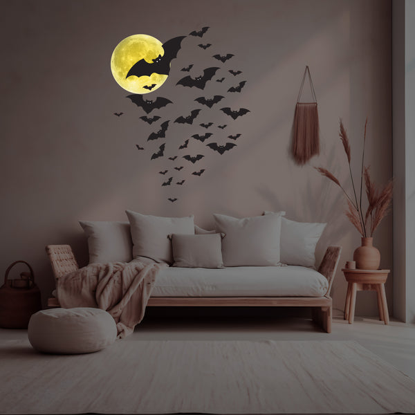 A minimalist living room with a white couch, beige pillows, and a throw blanket. The wall features a glowing Cover-Alls Silver or Yellow Moon Decal and bat decals, adding a celestial touch. A pouf, basket, side table, and wall hanging are also visible.