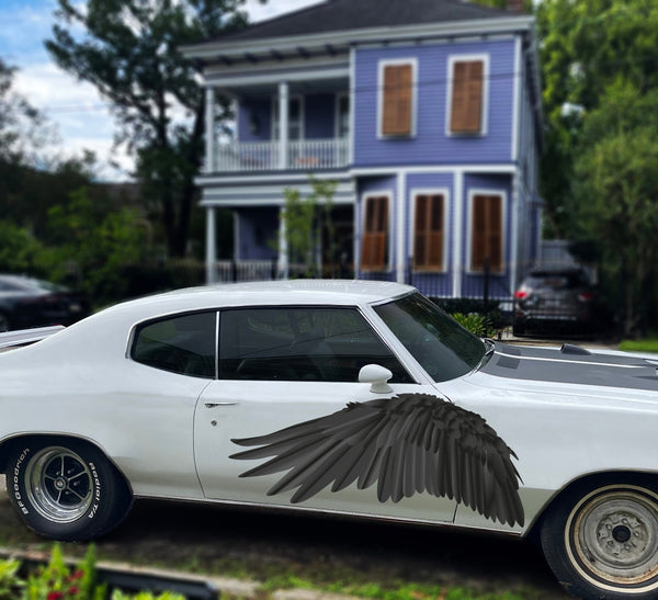 A white vintage car with large Cover-Alls black wings attached to the side, parked in front of a two-story purple house with greenery around.