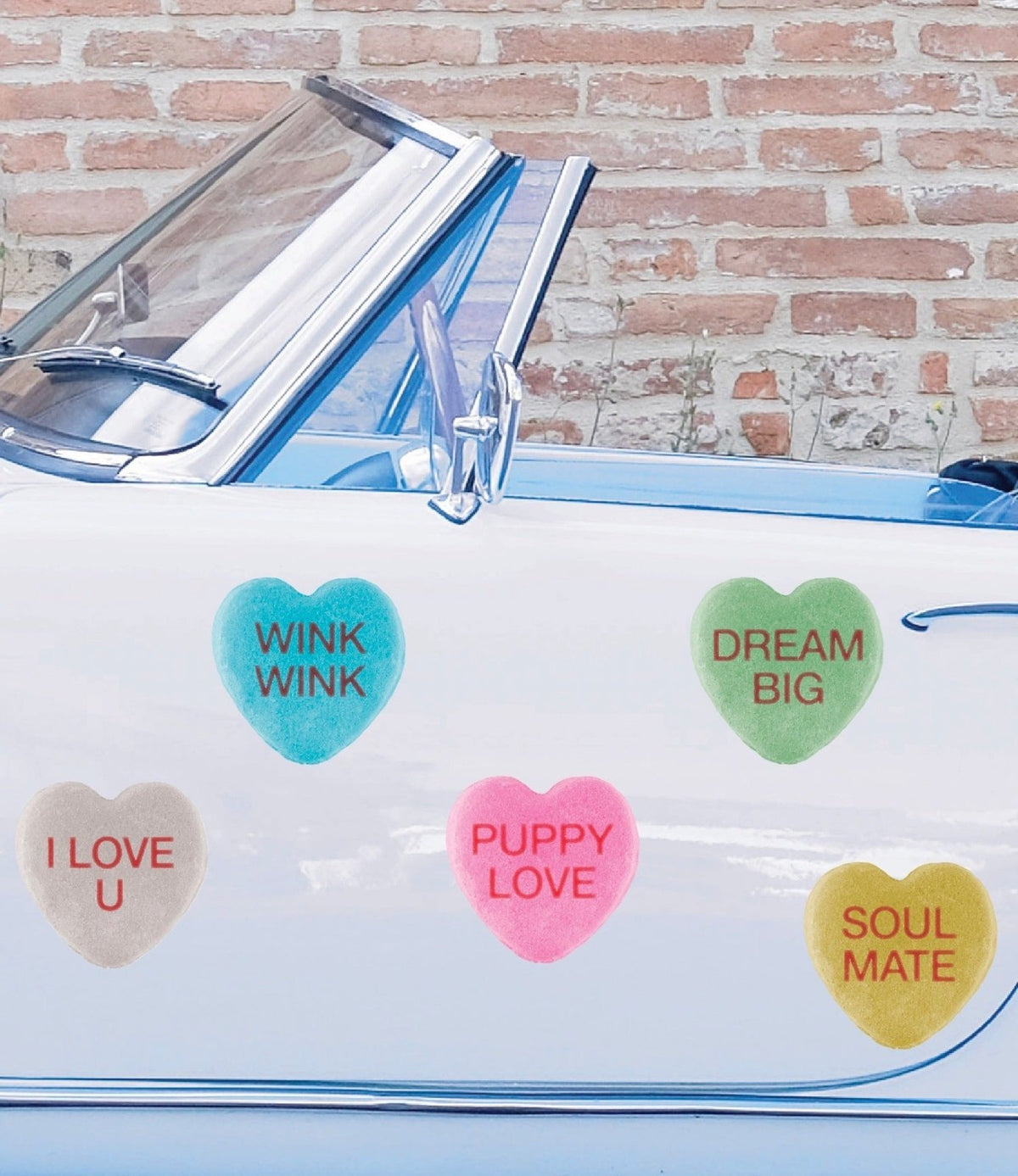 A vintage blue convertible car with heart-shaped stickers on the windshield displaying phrases like "wink wink" and "dream big" against a brick wall background, adorned with Cover-Alls candy hearts for Valentine's Day.