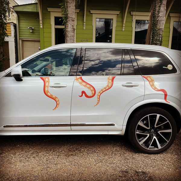 A white SUV with red flame decals and Eight Terrifying Tentacle Decals as a car decoration parked in front of a yellow building with green trim.