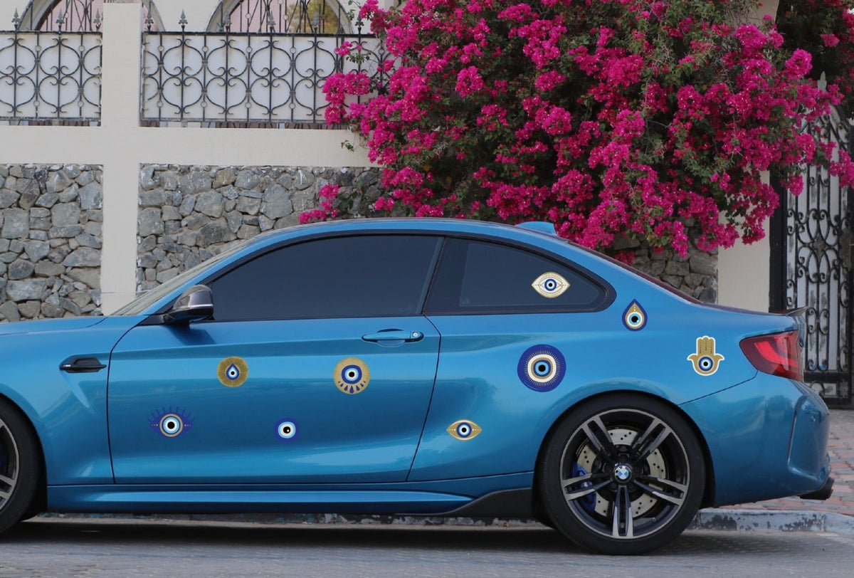 Blue car adorned with CoverAlls Evil Eye decals parked near a flowering bush.