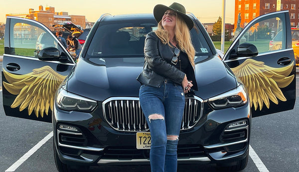 Woman in a black leather jacket and ripped jeans standing in front of a black SUV with Cover-Alls Gold Wing Decals for doors open.