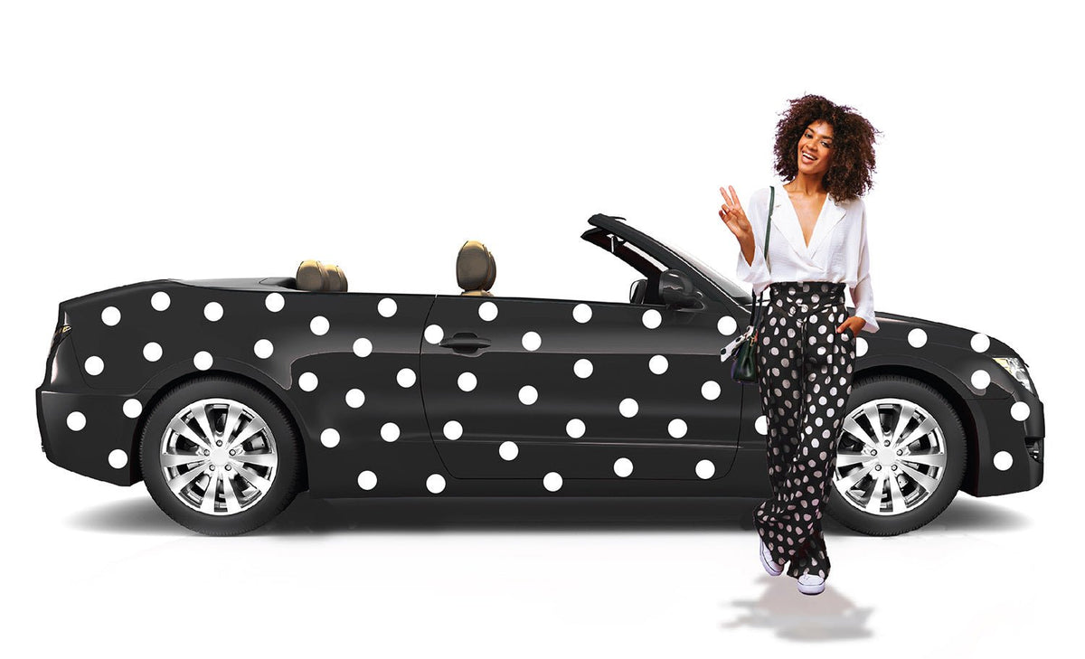 A woman with curly hair, standing by a black convertible car with Cover-Alls white polka dot decals, is smiling and waving. She wears a white blouse and polka-dotted pants.
