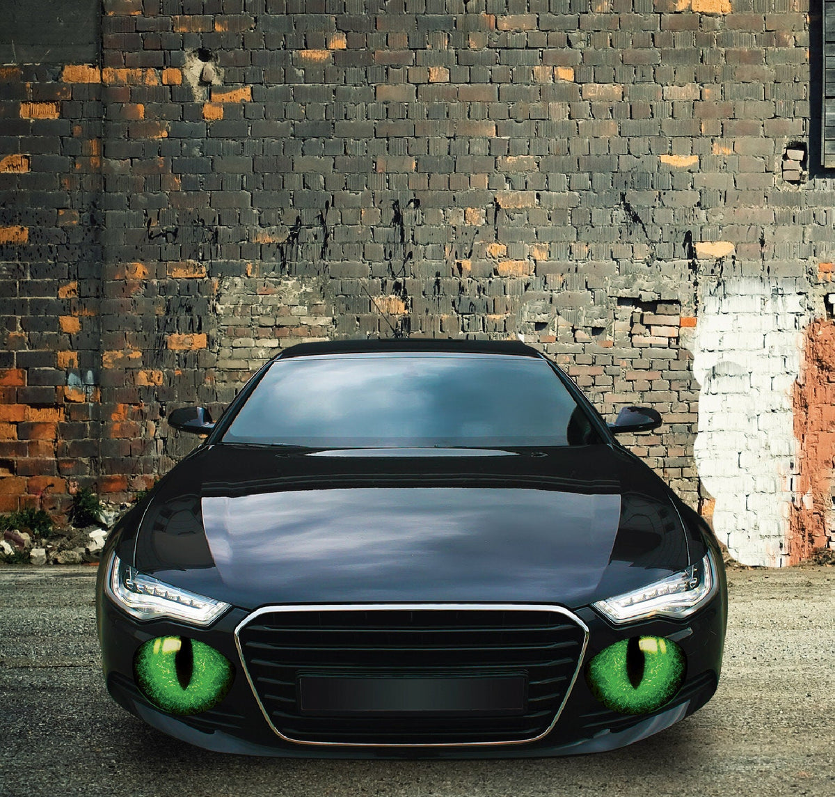 A black car with glowing green Cover-Alls cat eye decals in the grille, parked in front of a dark brick wall in a quiet neighborhood, with scattered white patches.